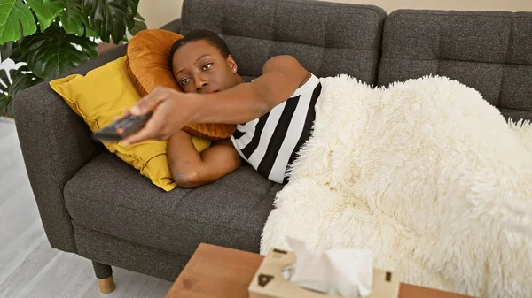 Exhausted african american woman sadly engrossed in a boring film, lying comfortably on the sofa, covering with a blanket, relaxing at her beautiful home interior in the living room