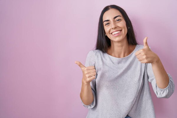 Young brunette woman standing over pink background success sign doing positive gesture with hand, thumbs up smiling and happy. cheerful expression and winner gesture. 