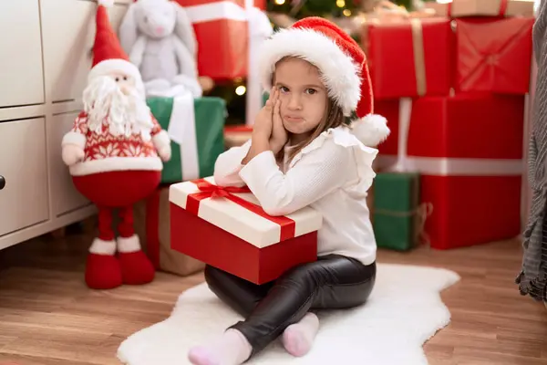愛らしいですヒスパニック女の子保持ギフトによって床に座ってクリスマスツリーとともに悲しい表現で家 ストック画像