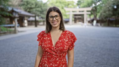 Cheerful, beautiful hispanic woman with glasses poses confidently, smiling at tokyo's meiji shrine, her joy unequivocally radiating through her astonishing smile clipart