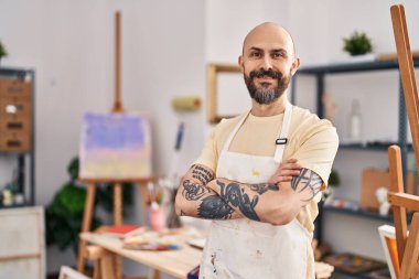 Genç kel adam gülümsüyor. Kollarını kavuşturup sanat stüdyosunda duruyor.