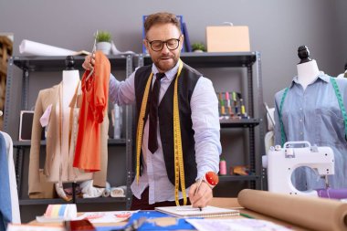 Middle age man tailor holding t shirt writing on notebook at atelier