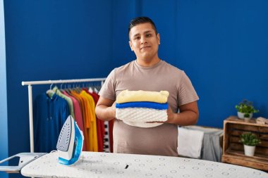 Yüzünde ciddi bir ifadeyle ütüledikten sonra katlanmış çamaşırları tutan İspanyol genç adam. Basit ve doğal bir şekilde kameraya bakıyorsun.. 