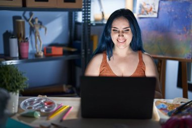 Mavi saçlı genç modern kız gece yarısı dizüstü bilgisayarla resim stüdyosunda oturuyor ve komik bir ifadeyle dilini dışarı çıkarıyor. duygu kavramı. 