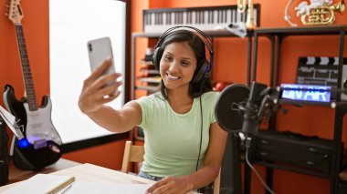 Kulaklık takan genç Latin kadın müzik stüdyosunda akıllı telefondan selfie çekiyor.