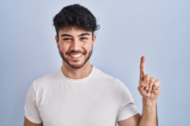 Hispanic man with beard standing over white background showing and pointing up with finger number one while smiling confident and happy. 