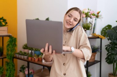 Young caucasian woman florist talking on smartphone using laptop at flower shop