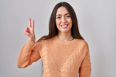 Genç esmer kadın beyaz arka planda duruyor. İki numaralı parmaklarıyla işaret ediyor ve gülümsüyor. Kendinden emin ve mutlu.. 