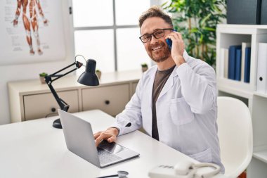 Kafkasyalı genç doktor laptopunu kullanarak klinikte akıllı telefondan konuşuyor.