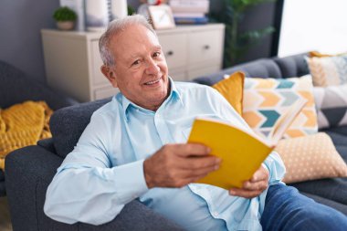 Orta yaşlı, kır saçlı, kitap okuyan adam evdeki kanepede oturuyor.