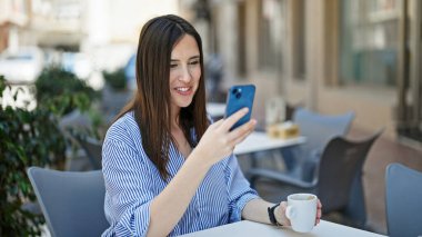 Young beautiful hispanic woman using smartphone drinking coffee at coffee shop terrace