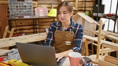 Ahşap atölyesinde dizüstü bilgisayarı ustalıkla kullanan, marangozluk işinde başarılı, gülümseyen sarışın kadın..