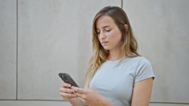 Neşeli genç sarışın kadın şehir sokaklarında akıllı telefonuyla eğlenceli bir selfie çekiyor. Şehir hayatının tadını çıkarırken gün ışığında güneşleniyor..