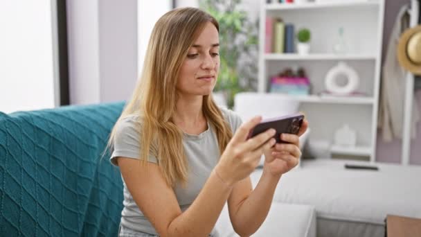 エキサイティングな若いブロンドの女性 彼女の快適なアパートで 快適に彼女のソファに座っている間応援 彼女のモバイルガジェットでエキサイティングなオンラインビデオゲームを再生することに興奮 — ストック動画