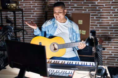 Latin asıllı genç bir adam müzik stüdyosunda klasik gitar çalıyor neşeli bir şekilde sunum yapıyor ve avuç içi kameraya bakıyor.. 
