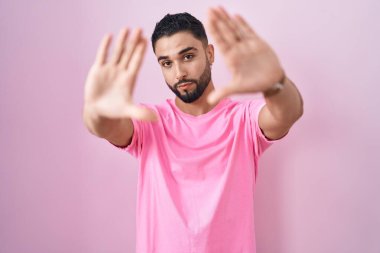 İspanyol genç adam pembe arka planda duruyor. El ve parmakları kullanarak çerçeve yapıyor. Kamera bakış açısı. 