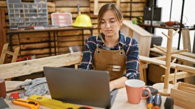 Ciddi sarışın marangoz kadın ev içi marangozluk atölyesinde online ahşap işi için dizüstü bilgisayar kullanıyor.