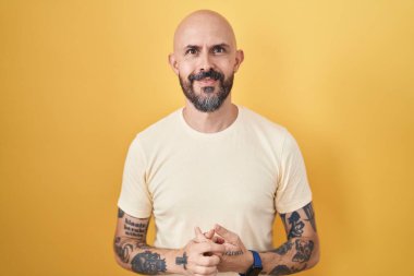 Sarı arka planda dikilen İspanyol dövmeli adam ellerini kavuşturup gülümseyerek gevşemiş ve neşeliydi. Başarı ve iyimserlik 