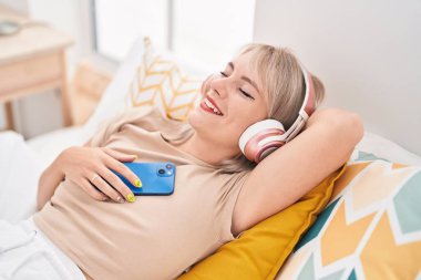 Young blonde woman listening to music relaxed on bed at bedroom