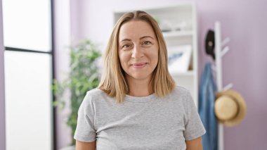 Neşeli genç sarışın kadın, evin olumlu havasının tadını çıkarıyor, kendinden emin gülüşü neşe saçıyor, güzel apartman odasının sıcak, kapalı kapılar ardında duruyor..