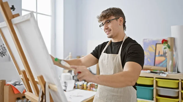 Cheerful young hispanic man, an artist enwrapped in passion, confidently drawing in art studio with a smile