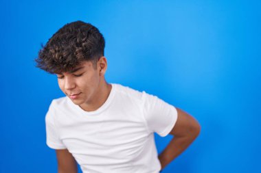 Mavi arka planda duran İspanyol ergen sırt ağrısı çekiyor, elleriyle dokunuyor, kas ağrısı çekiyor. 