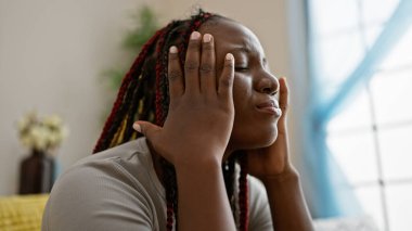 Üzüntülü, örgülü, evde tek başına oturan Afro-Amerikan kadın. Mutsuz yüz ifadesi baş ağrısı ve stresi gösteriyor.