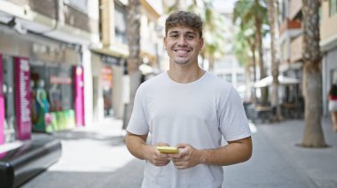 Genç İspanyol adam sokakta gülümseyen akıllı telefon kullanıyor.