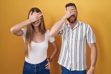 Sarı arka planda dikilen genç bir çift. Gülümsüyor ve gülümsüyor. Gözleri şaşkınlıkla örtüyor. kör kavram. 