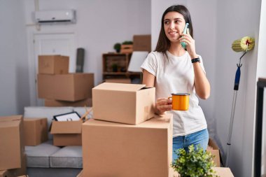 Young beautiful hispanic woman talking on smartphone drinking coffee at new home