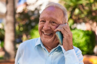 Orta yaşlı, kır saçlı, gülümseyen, kendine güvenen, parkta akıllı telefondan konuşan bir adam.