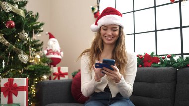 Genç ve güzel İspanyol kadın akıllı telefon ve kredi kartı kullanıyor. Evdeki Noel ağacının yanında, koltukta oturuyor.