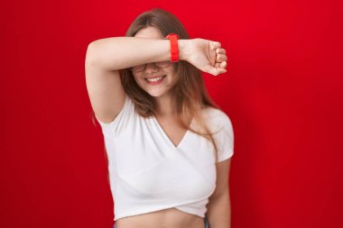 Genç beyaz kadın kırmızı arka planda duruyor. Gözleri kollarıyla kaplıyor. Neşeli ve komik gülümsüyor. kör kavram. 