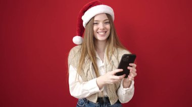 Yılbaşı şapkası takan genç sarışın kadın izole edilmiş kırmızı arka plan üzerinde akıllı telefon kullanıyor.