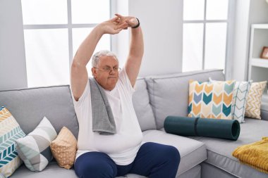 Orta yaşlı, kır saçlı, kollarını esneten adam evdeki kanepede oturuyor.