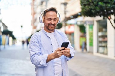 Genç, beyaz bir adam gülerek sokakta müzik dinliyor.