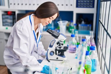 Young beautiful hispanic woman scientist using microscope writing on document at laboratory