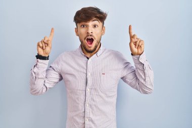 Mavi arka planda duran sakallı Arap adam şaşırmış, şaşırmış ve parmaklarıyla işaret ediyor ve kollarını kaldırıyordu.. 