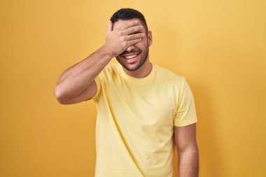 Young hispanic man standing over yellow background smiling and laughing with hand on face covering eyes for surprise. blind concept. 