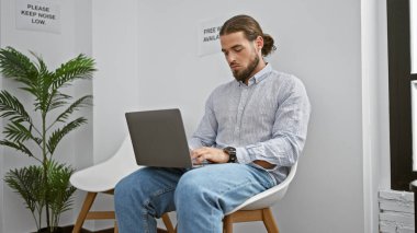 Genç İspanyol adam bekleme odasında otururken dizüstü bilgisayar kullanıyor.