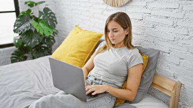 Yatak odasında dizüstü bilgisayar kullanan genç sarışın kadın.