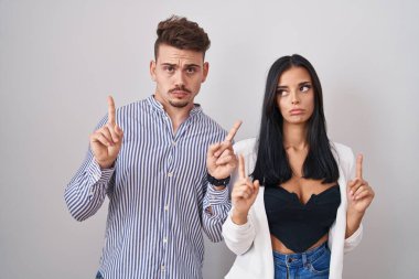 Young hispanic couple standing over white background pointing up looking sad and upset, indicating direction with fingers, unhappy and depressed. 