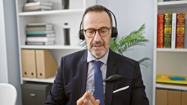 Ofiste konuşan, gri saçlı, kulaklık takan orta yaşlı bir iş adamı.