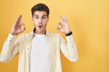 Young hispanic man standing over yellow background looking surprised and shocked doing ok approval symbol with fingers. crazy expression 