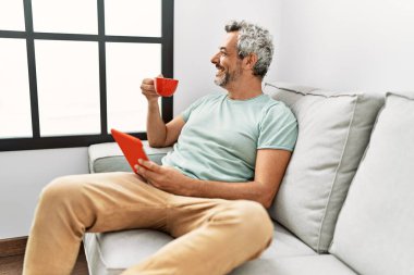 Middle age grey-haired man using touchpad drinking coffee sitting on sofa at home
