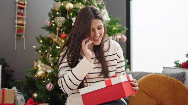 Genç, güzel, İspanyol bir kadın elinde noel hediyesiyle evde akıllı telefondan konuşuyor.