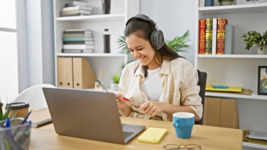Neşeli genç İspanyol kadın işçi, ofiste laptopuyla çalışırken müzik eşliğinde dans ediyor, kendine güveni tam.