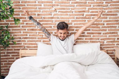 Young hispanic man waking up stretching arms at bedroom