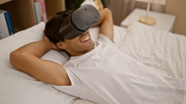 Çevrimiçi oyundan büyülenmiş mutlu İspanyol genç adam, fütüristik VR kulaklığın tadını çıkarıyor, elinde aletle yatak odasında yatıyor, oyun yeteneğine güveniyor, göz alıcı bir gülümseme ile kapalı alanda hevesli..