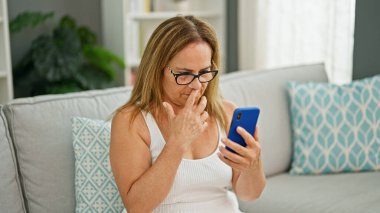 Orta yaşlı İspanyol kadın kanepeye oturmuş akıllı telefon kullanıyor. Evinde üzgün görünüyor.